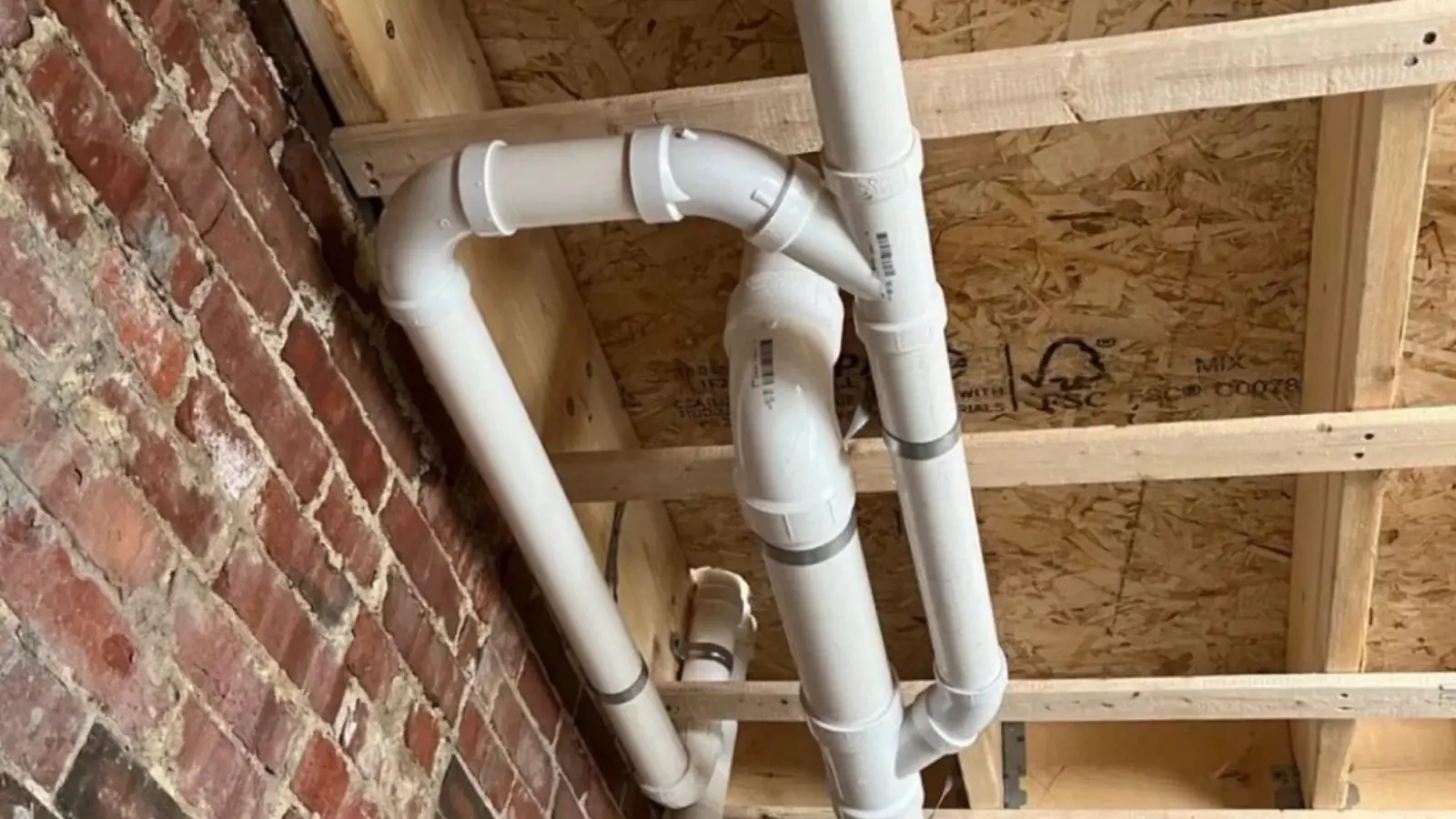 New white PVC drain piping running past an old red brick basement wall and wood framing