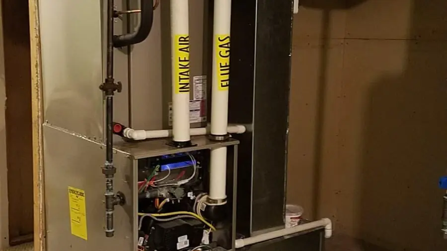 A high-efficiency gas furnace with its front panel off, showing the inside of the unit, with white PVC pipes labelled intake air and flue gas running above it