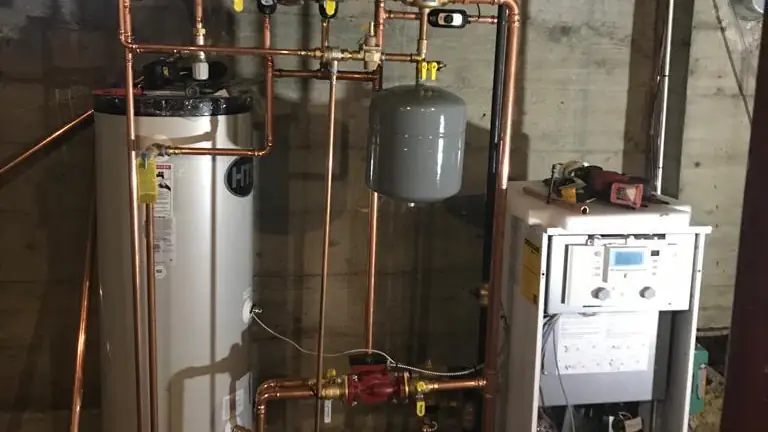 A tall stainless hot water storage tank beside a boiler with copper piping and an expansion tank in a basement