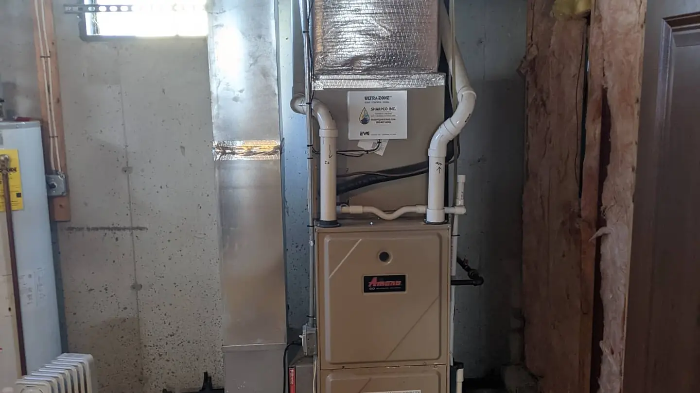 Gas furnace with white PVC vent pipes and metal ductwork in an unfinished basement, with an old radiator on the left