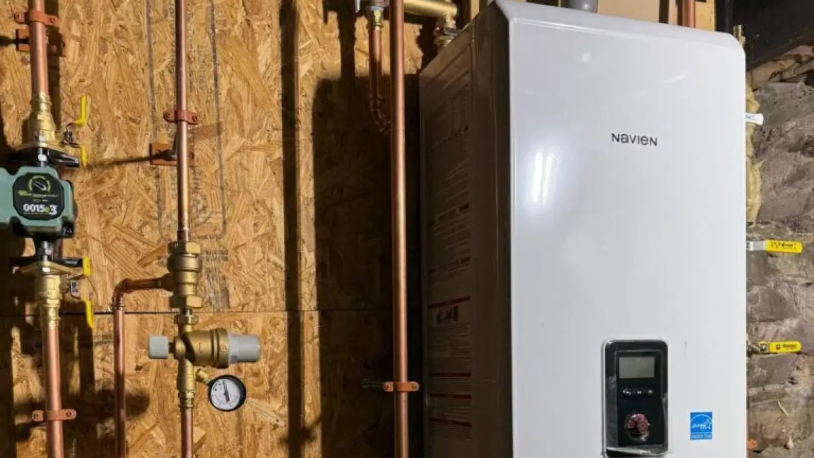 A white wall-mounted combi boiler with a digital display, copper piping and a circulator pump on a plywood wall