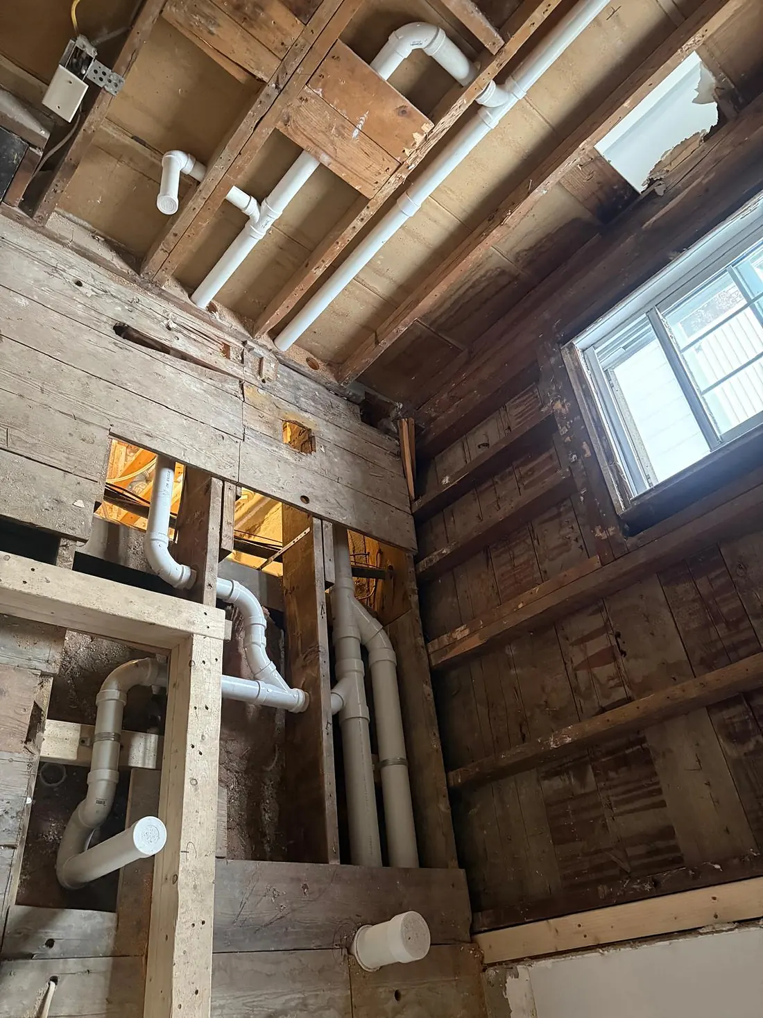Basement under renovation with open wood framing and exposed joists, showing new white plastic drain and vent piping running through the walls and ceiling beside a window