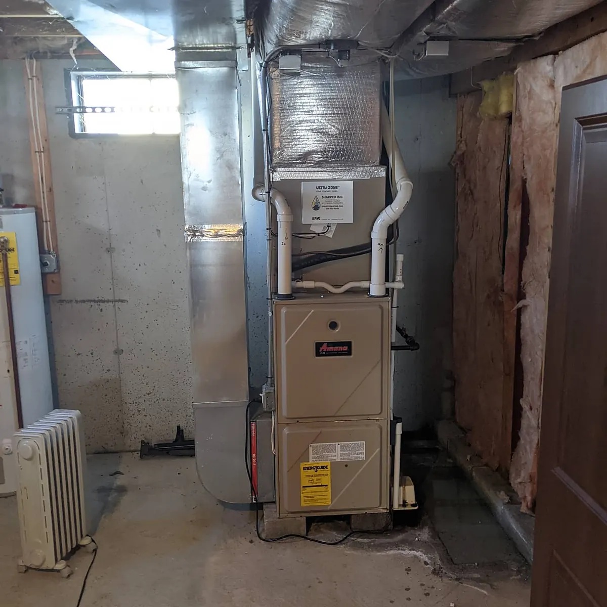 Gas furnace with white PVC vent pipes and silver ductwork in an unfinished concrete basement, with an electric water heater and a portable radiator nearby