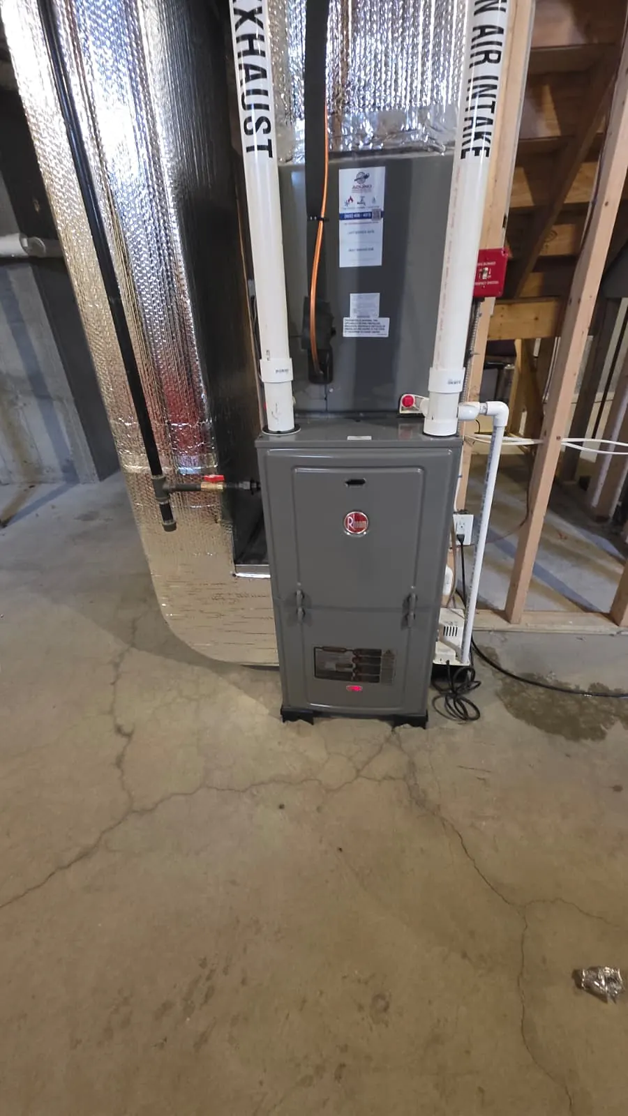 Grey high-efficiency gas furnace in an unfinished basement with two white PVC pipes above it labeled exhaust and air intake, next to an insulated tank and wood stair framing